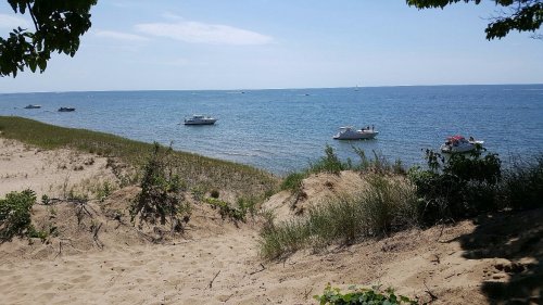 A serene beach scene with boats on calm water, surrounded by sandy dunes and green grass under a clear blue sky.