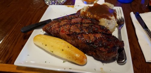 A plate with a grilled steak, garlic bread, and mashed potatoes topped with gravy, set on a wooden table.
