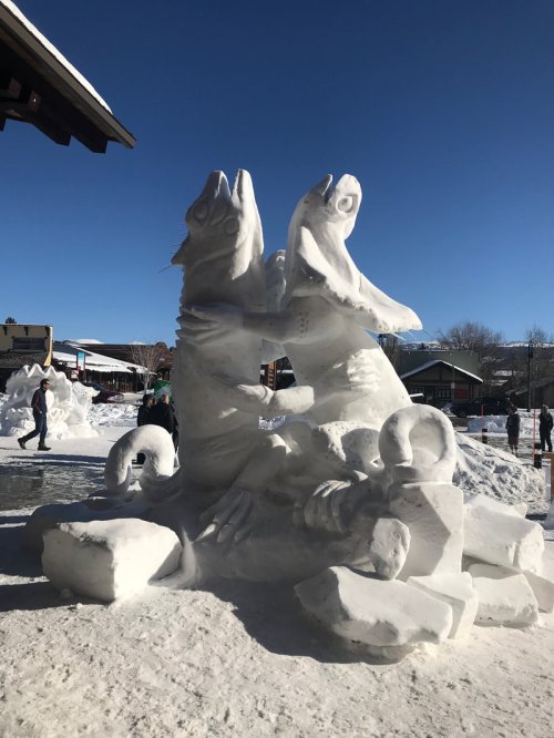 A large snow sculpture of two fish facing each other, surrounded by snow and people in a winter setting.