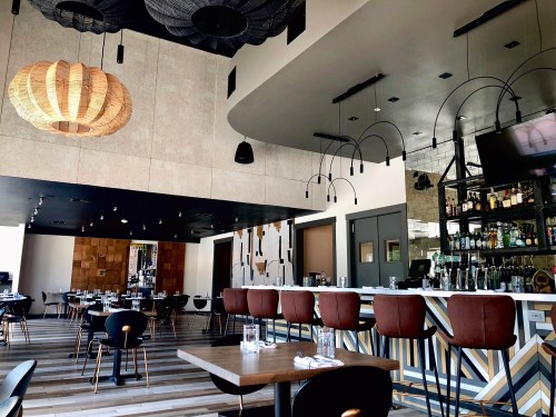 Modern restaurant interior featuring a bar, stylish lighting, and wooden tables with black chairs.