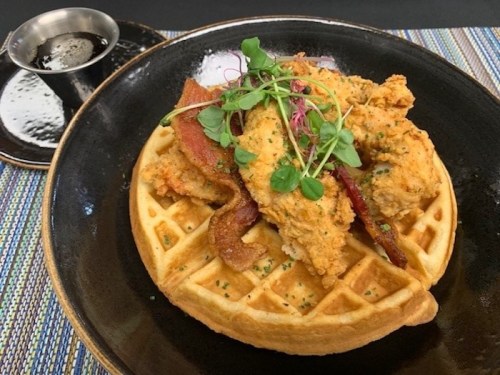 A plate of fried chicken and bacon atop a golden waffle, garnished with microgreens, served with a side of syrup.