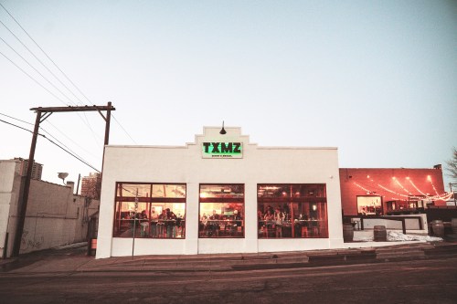 A white restaurant building with large windows, featuring a green sign that reads "TXMZ," set against a twilight sky.