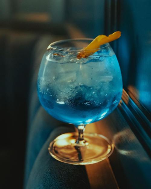 A blue cocktail with ice and a twist of orange peel, served in a round glass on a table.