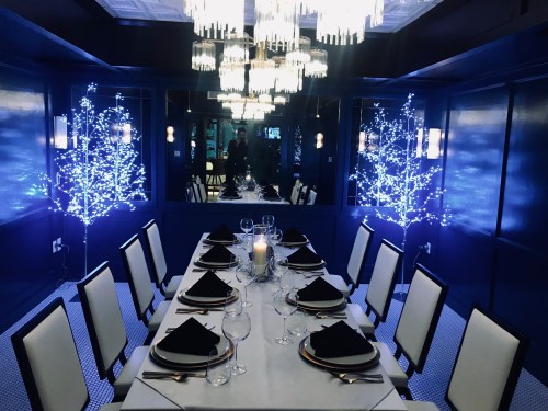 Elegant dining room with a long table set for dinner, surrounded by glowing blue trees and a sparkling chandelier.