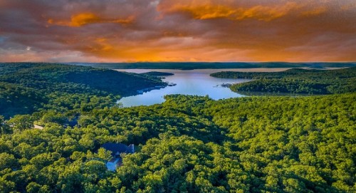 Aerial view of a lush green landscape with a lake under a dramatic orange and purple sunset sky.
