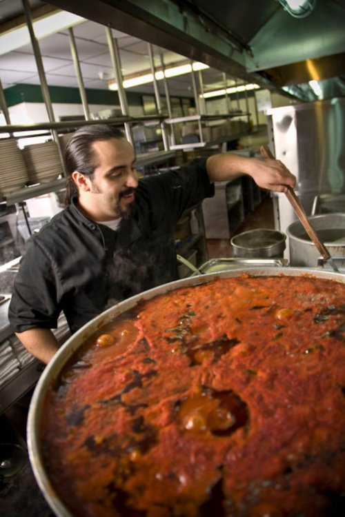 A chef stirs a large pot of simmering red sauce in a busy kitchen. Steam rises around him.