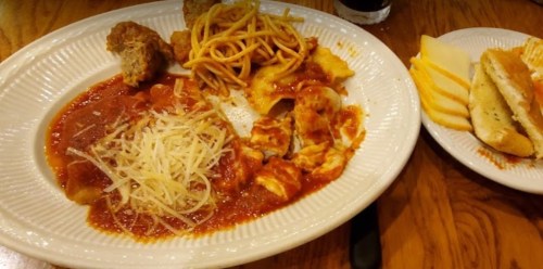 A plate of pasta with marinara sauce, cheese, breadsticks, and meatballs on a wooden table.