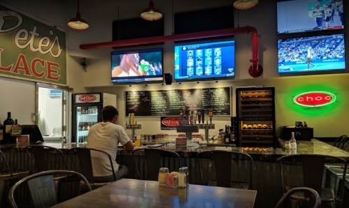 A bar with a man sitting alone, multiple TVs showing sports, and a neon sign reading "Choc." Cozy atmosphere.