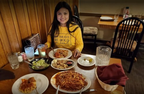 A woman sits at a table with a variety of dishes, including spaghetti, salad, and chicken, in a cozy restaurant setting.