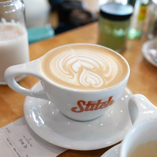 A latte with intricate foam art in a white cup on a wooden table, with blurred background items.
