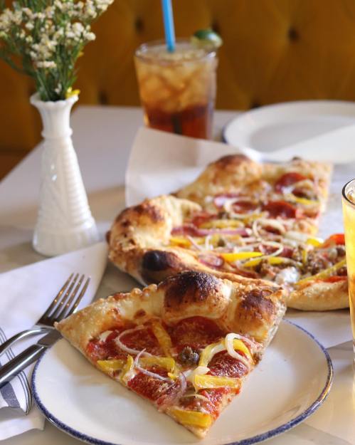 Two slices of pepperoni pizza topped with onions and peppers, served on white plates with drinks in the background.