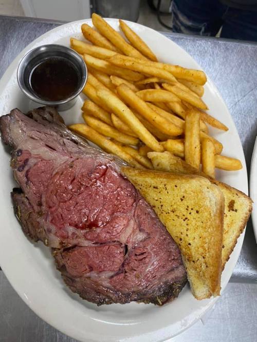 A plate featuring a large slice of prime rib, crispy fries, a piece of toast, and a small cup of sauce.