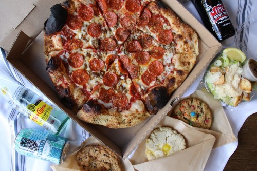 A pizza with pepperoni and sausage, cookies, salad, and drinks arranged in a takeout box on a table.