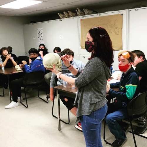 A teacher holds a skull while students observe in a classroom setting, all wearing masks.