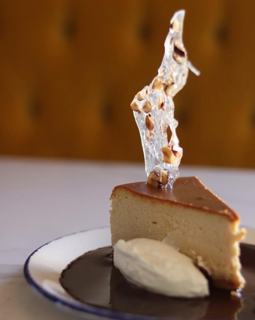 A slice of cheesecake topped with a caramel shard and nuts, served with a dollop of whipped cream on a plate.