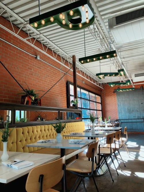 Bright, modern restaurant interior with brick walls, yellow tufted seating, and hanging lights. Tables set for dining.