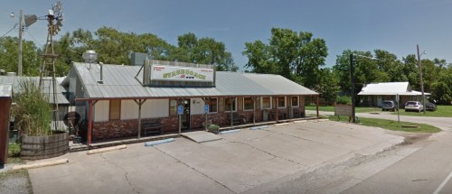 A rustic restaurant with a metal roof and stone accents, surrounded by trees and a parking area.