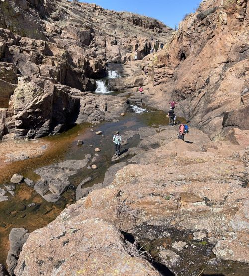Hikers navigate rocky terrain near a stream and waterfall in a rugged landscape.