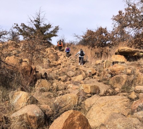 Three hikers navigate a rocky trail surrounded by sparse trees and dry grass under a cloudy sky.