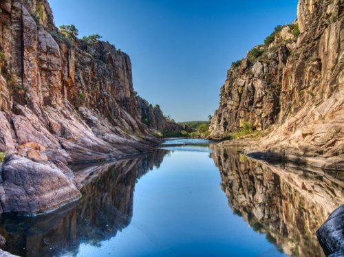 A serene river flows between steep, rocky cliffs under a clear blue sky, reflecting the landscape in calm waters.