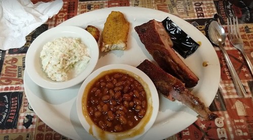 A plate of barbecue ribs, cornbread, coleslaw, baked beans, and potato wedges on a patterned tablecloth.