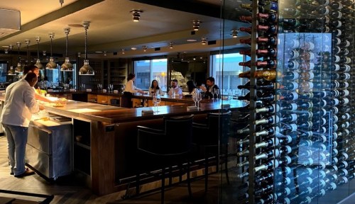A modern restaurant interior featuring a bar, wine display, and chefs preparing food in a stylish setting.