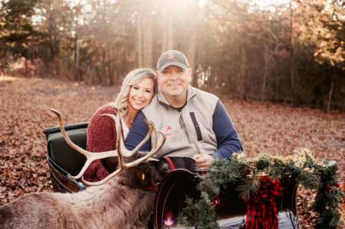 A couple smiles together in a sleigh surrounded by autumn leaves, with a reindeer and festive decorations nearby.