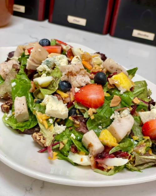 A colorful salad with greens, chicken, strawberries, blueberries, cheese, and nuts, served on a white plate.