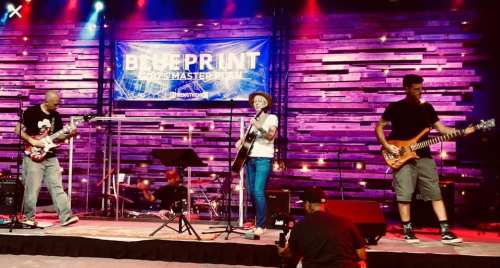 A band performs on stage with guitars and drums, illuminated by colorful lights and a backdrop banner.
