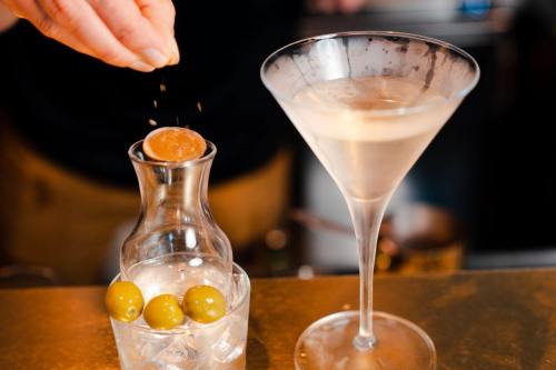A hand sprinkles seasoning over a cocktail glass next to a carafe of olives and ice.