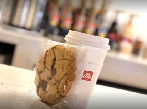 A coffee cup next to a chocolate chip cookie, both on a marble countertop with blurred background items.