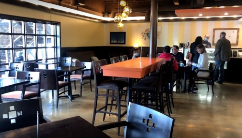 A cozy café interior with wooden tables, chairs, and a vibrant orange bar, featuring a few patrons enjoying their time.