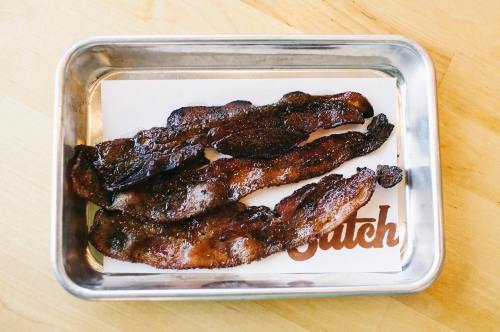 A metal tray holding several pieces of crispy, dark-brown bacon on a branded paper liner.