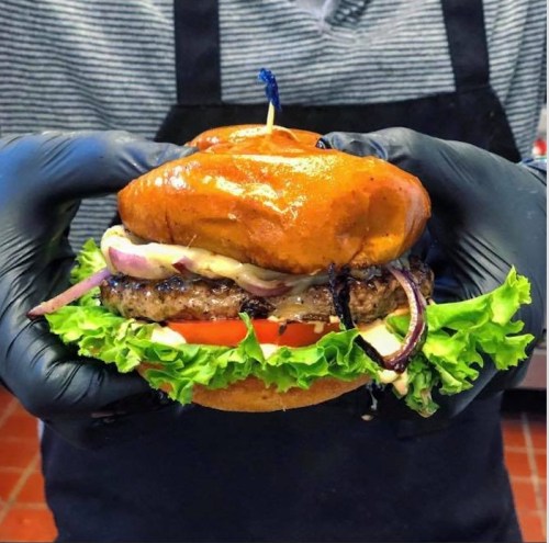 A person in black gloves holds a juicy burger with lettuce, tomato, onions, and a shiny bun, topped with a toothpick.