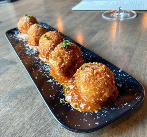 A black plate with five golden, crispy balls drizzled with sauce and garnished with herbs on a wooden table.