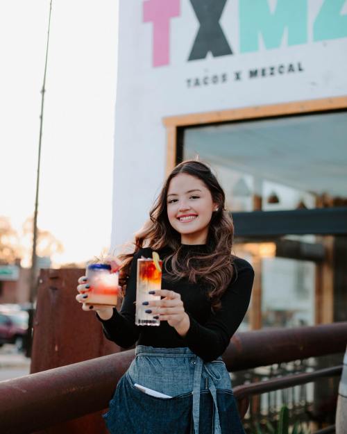 A smiling woman holds two colorful cocktails outside a restaurant with a sign that reads "Tacos x Mezcal."