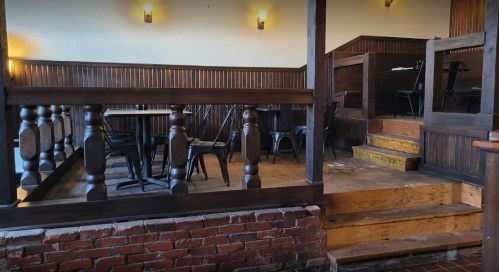 Interior of a rustic restaurant with wooden decor, empty tables, and a small raised area with stairs.