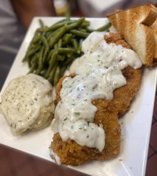 A plate with fried chicken covered in white gravy, green beans, mashed potatoes, and a slice of toast.