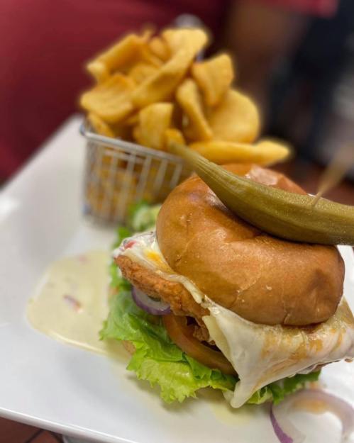A close-up of a burger with lettuce, tomato, and a fried egg, served with crispy potato chips and a pickle on top.