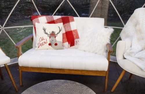 Cozy seating area with a white couch, red plaid blanket, and festive pillow in a geodesic dome.