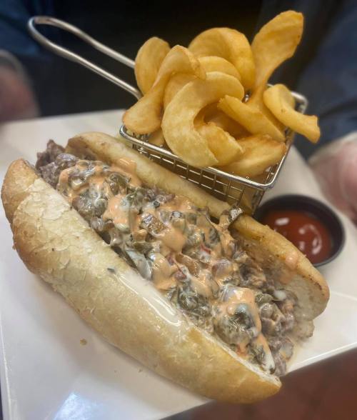 A loaded sandwich with beef, toppings, and sauce, served with a side of curly fries and ketchup.