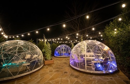 Three illuminated geodesic domes in a garden, surrounded by string lights and greenery at night.