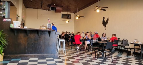 A spacious café with checkered floors, featuring patrons seated at tables and a large rooster mural on the wall.