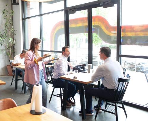 A waitress serves customers at a modern café with large windows and colorful decor.