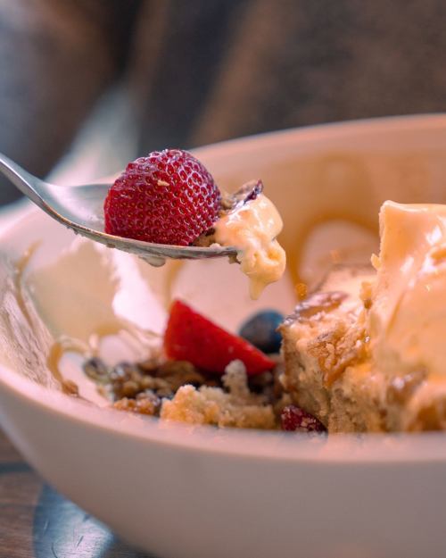 A spoon lifts a strawberry and creamy dessert from a bowl, with crumbs and berries visible in the dish.