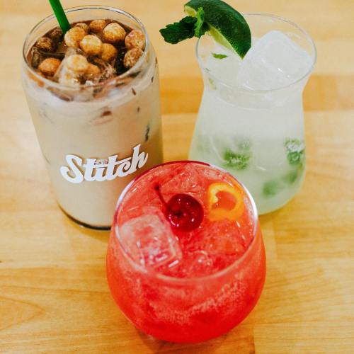 Three colorful drinks on a wooden surface: a creamy iced coffee, a minty green cocktail, and a vibrant red beverage with a cherry.