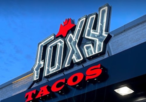 Neon sign for "Foxy Tacos" illuminated against a twilight sky.