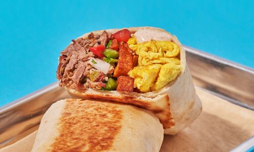 A close-up of a burrito filled with beef, eggs, tomatoes, and peppers, resting on a tray against a blue background.
