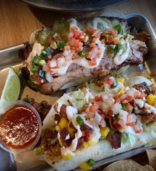 Two loaded tacos topped with lettuce, pico de gallo, and sauces, served with lime and a side of salsa.