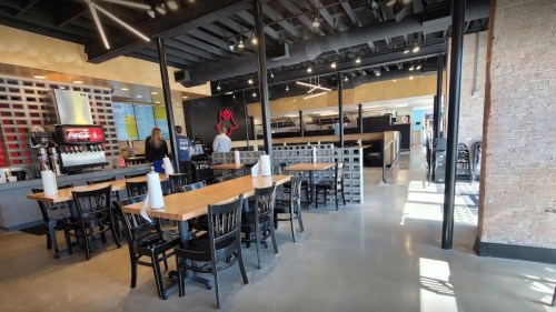 Interior of a modern restaurant with wooden tables, black chairs, and a soda fountain, featuring a bright, open layout.